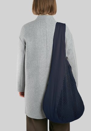 Person in einem hellgrauen Mantel und dunklen Hosen, der eine große marineblaue Netz-Tote-Tasche über die Schulter trägt, vor einem schlichten Hintergrund.