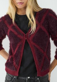 Bordowy, puszysty kardigan z diamentowym wzorem; miękka tekstura, długie rękawy i zapięcie na guziki. Noszony na czarnej koszulce i ciemnych dżinsach.