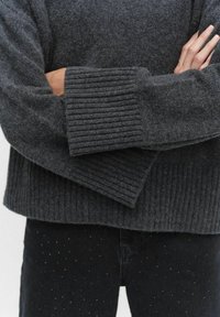 Donkergrijze trui gemaakt van een zachte, gestructureerde stof, met brede ribboorden en een losse pasvorm. Gecombineerd met zwarte jeans versierd met kleine studs.