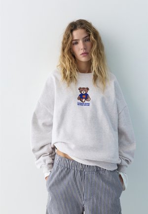 Jeune femme aux cheveux blonds ondulés portant un sweat-shirt gris clair avec un motif d'ourson et un pantalon rayé bleu et blanc, les mains dans les poches.
