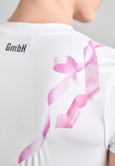T-shirt blanc avec un grand motif de ruban rose au dos, d'une texture lisse et un logo noir "GmbH" au niveau du col.