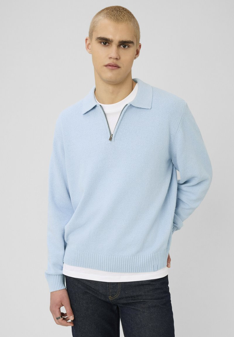 Jeune homme aux cheveux blonds courts, portant un pull à col zippé bleu clair par-dessus une chemise blanche et un jean foncé, debout devant un fond gris uni.
