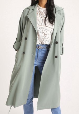 Femme portant un long manteau croisé vert clair sur un haut en dentelle blanche et un jean bleu avec une ceinture chaîne dorée, mains dans les poches du manteau.