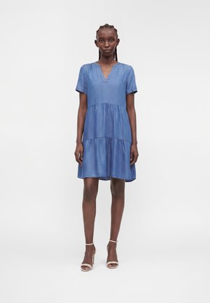 Mujer de pie, mirando hacia adelante, vistiendo un vestido azul con volantes y mangas cortas y sandalias blancas de tacón alto sobre un fondo liso.