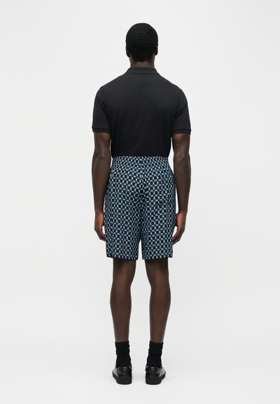 Άνδρας που φοράει μαύρο πουκάμισο με κοντά μανίκια και μπλε-μαύρο βερμούδα με σχέδια μέχρι το γόνατο, στέκεται γυρισμένος προς τα πίσω μπροστά σε απλό φόντο.