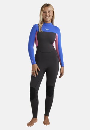 Frau steht barfuß, trägt einen schwarzen Neoprenanzug mit blauen Schultern und pinken Seitenteilen, lächelt und hat die Hände entspannt an den Seiten.