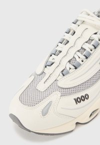 Chaussure de sport blanche et grise en mesh et matériau synthétique, dotée de détails texturés, d'un design de laçage unique et d'une décoration avec logo.