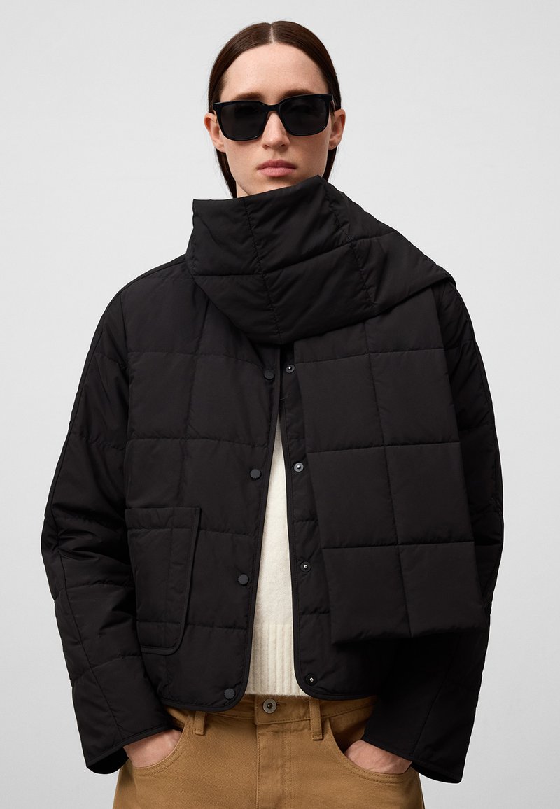 Giubbotto imbottito nero con colletto alto, dotato di bottoni automatici, tasca laterale e una texture morbida, indossato sopra un maglione beige.