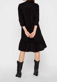 Mustapuku, jossa on liehuva helma, napitettu hihansuut ja poimutettu selkäosa. Yhdistettyinä korkeisiin mustiin saappaisiin, esittelee yksinkertaista, tekstuuripintaista kangasta.