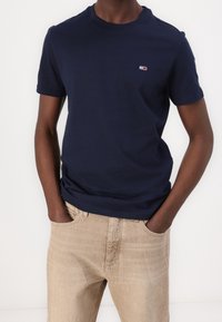 Camiseta de algodón azul marino con cuello redondo y mangas cortas, con un pequeño logo bordado en el pecho izquierdo. Combinada con vaqueros beige.