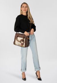 Braune Handtasche mit einem dekorativen Frontpaneel, das eine Mädchenillustration und Text zeigt. Verfügt über zwei Griffe und einen Schultergurt.