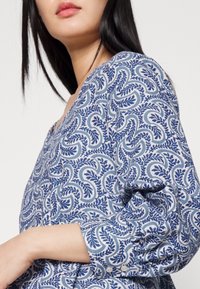Blaue Bluse mit gemustertem botanischem Design, puffärmeligen Ärmeln und Knopfdetails an den Bündchen. Glatte Stofftextur.