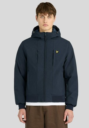 Jonge man draagt een marineblauwe capuchonjas met een geel vogel-logo, wit onderhemd en een bruine broek, staand tegen een lichtgrijze achtergrond.