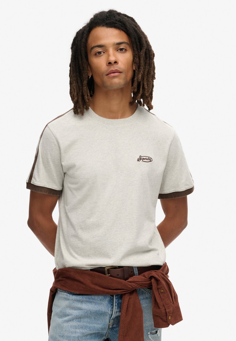 Lysegrå t-shirt med brune ærmer og kravekant, broderet "Superdry" logo på brystet. Kombineret med blå denimjeans og et brunt kordbælte.