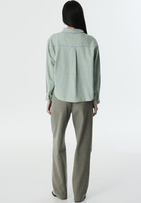 Camicia di denim verde chiaro con orlo curvo e piega sul retro, abbinata a pantaloni grigi larghi e testurizzati con tasche posteriori.
