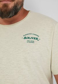 Beige T-Shirt mit kurzen Ärmeln und rundem Ausschnitt. Mit grünem Drucktext "S. OLIVER" und zusätzlichen Markendetails. Weicher Stoff.