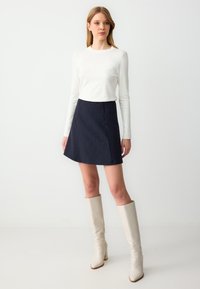 Weißes Langarmoberteil kombiniert mit einem mini Rock in Marineblau mit Nadelstreifen. Das Outfit wird durch cremefarbene kniehohe Stiefel ergänzt. Glatte Texturen.