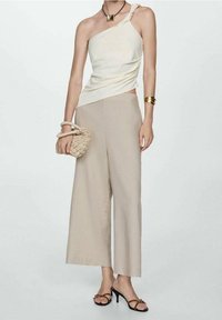 Žena oblečená v krémovém topu na jedno rameno, béžových širokých kalhotách, černých sandálech na podpatku, zlatém náramku a držící malou tkánou kabelku.
