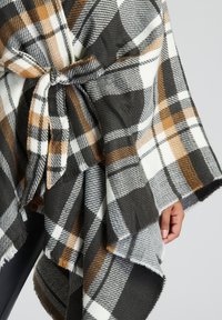 Rutig poncho i svart, vitt och orange, med knytbar midja, mjuk textur och fransade kanter för extra detalj.