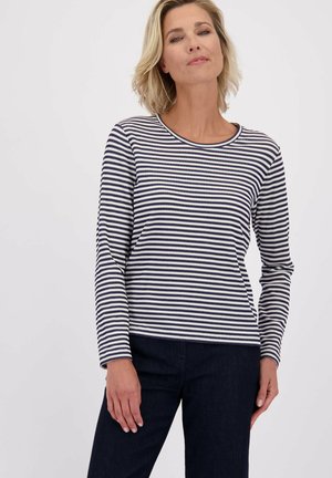 Vrouw met kort blond haar, gekleed in een langwerpige, marineblauwe en witte horizontaal gestreepte shirt met lange mouwen, en donkere broeken tegen een effen achtergrond.