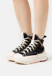 Sneaker alte in tela nera con lacci bianchi, occhielli dorati e suola spessa di colore crema. Presenta un accento a catena sul lato.
