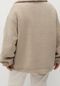 Beige fleecejakke med en struktureret overflade, oversized design, brede ærmer og krave. Sømdetaljer løber lodret ned ad ryggen.