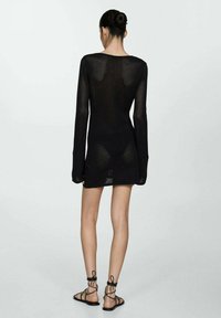 O femeie întoarsă, purtând o rochie lungă din tricot transparent negru cu mâneci lungi și sandale plate negre cu șireturi, pe un fundal simplu.