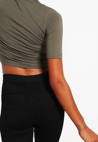 Haut court vert olive à manches courtes, avec une texture froncée. Associé à un pantalon noir taille haute, vue de dos.