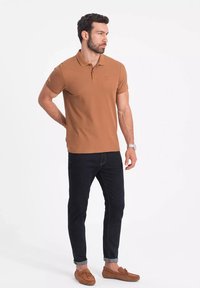 Brun polo-skjorta, kortärmad, liten logotyp på bröstet, i kombination med mörka jeans. Modell som bär bruna mockaskor och står avslappnat.