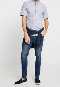 Camisa de manga corta azul claro con estampado y cierre de botones, jeans azul oscuro y zapatos náuticos azul marino. Suéter colocado alrededor de la cintura.
