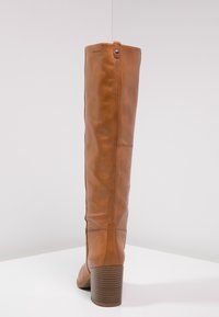 Botte en cuir marron, jusqu'au genou, avec un bout arrondi et un talon bloc texturé. Présente une couture latérale et un bouton décoratif en haut.