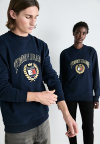 Sudadera de algodón azul marino con un escote redondo, que presenta un logo dorado y un diseño de laurel en el centro. Modelo vestido de manera casual.