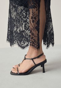 Sandales à talons noirs avec fines lanières et un design à bout carré. Le talon est de hauteur moyenne, complétant le tissu en dentelle noire délicate à proximité.