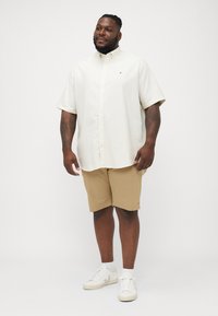 Short-sleeve, collared, cream shirt, paired with tan shorts and white sneakers. Features a small logo on the shirt. Smooth texture.
