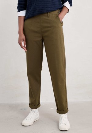 Personne portant un pantalon droit vert olive avec des revers et des baskets blanches, debout avec une main dans la poche et un pull bleu marine.
