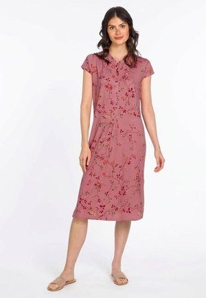 Vrouw die een roze bloemenjurk tot op de knie draagt met korte mouwen en beige sandalen met open tenen, staand tegen een witte achtergrond.