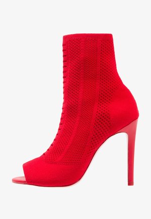 Czerwony botek za kostkę z odkrytymi palcami, wykonany z miękkiego materiału o teksturowanym, dzianinowym wzorze, z wysokim obcasem w szpilce i gładką, błyszczącą podeszwą.