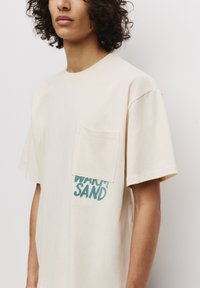Beige T-shirt med korte ærmer lavet af bomuld, med en frontlomme og turkis tekst der siger "WARM SAND" på venstre side.