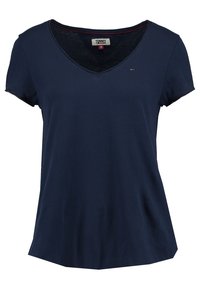 T-shirt en coton bleu marine avec un col en V, des manches courtes et une coupe décontractée. Présente des accents rouges sur le col et un petit emblème de logo.