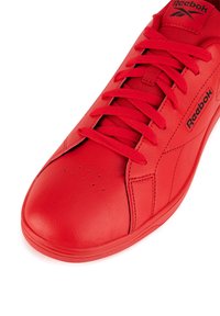 Raudona Reebok sportinė avalynė iš lygaus odos, apvali noselė ir plokščias padai; pasižymi ryškia raištelių sistema, logotipo detalėmis ir perforuotais akcentais.