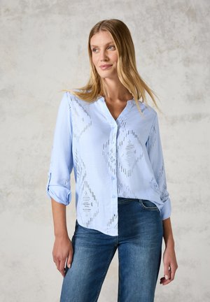 Lys blå skjorte med knaplukning og diagonale stribemønstre, lange ærmer oprevet til albuerne, iført mørke denimjeans.