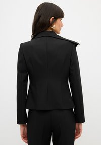 Blazer nero su misura con un design strutturato, dettagli di spalle affilate e texture liscia, abbinato a pantaloni eleganti aderenti.