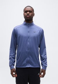 Άνδρας που φοράει ένα μπλε μπουφάν Nike με φερμουάρ και μαύρο παντελόνι Nike, στέκεται με τα χέρια χαλαρά μπροστά σε απλό λευκό φόντο.