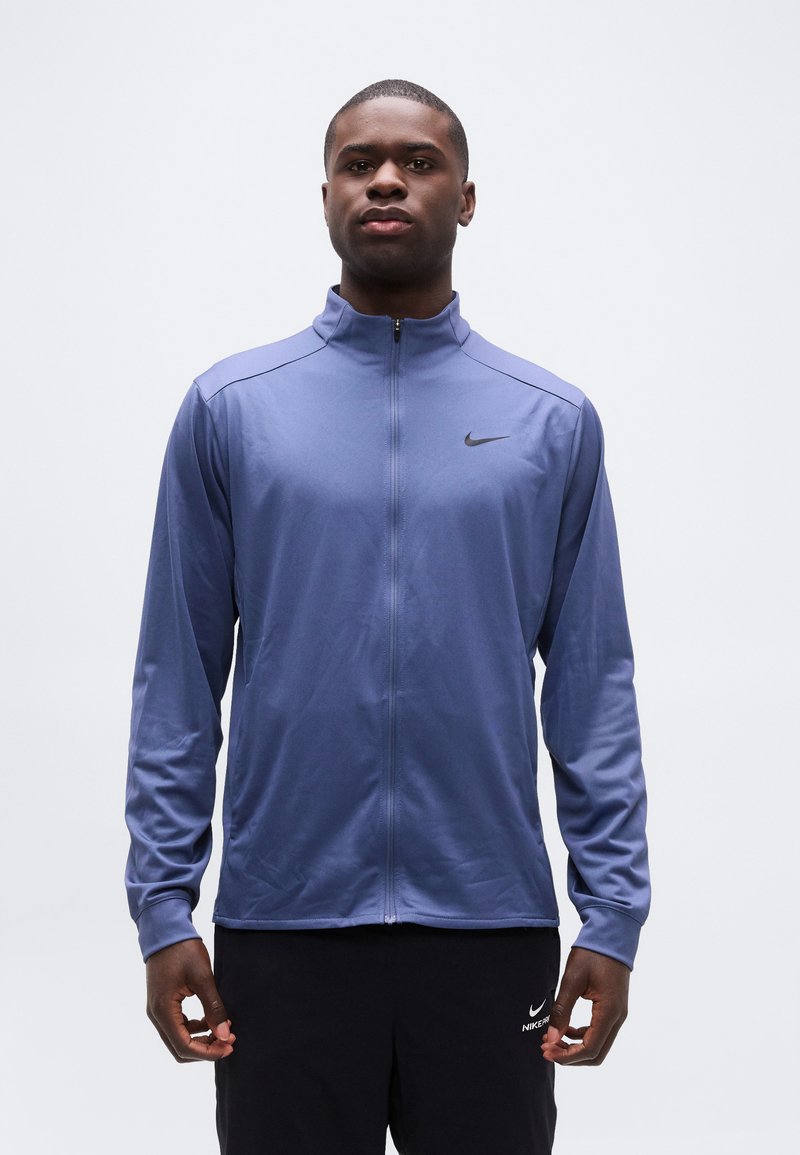 Άνδρας που φοράει ένα μπλε μπουφάν Nike με φερμουάρ και μαύρο παντελόνι Nike, στέκεται με τα χέρια χαλαρά μπροστά σε απλό λευκό φόντο.