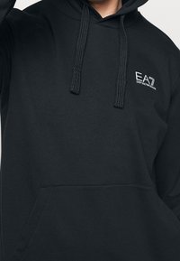 Černá mikina s kapucí a přední kapsou, na hrudi má bílé logo "EA7 Emporio Armani" a silné šňůrky na kapuci.