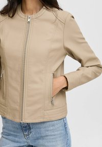 Chaqueta de cuero beige con cuello redondo, cierre frontal con cremallera, bolsillos laterales con cremallera, costuras acolchadas en las mangas y un silueta ajustada.