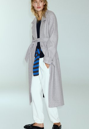 Femme aux cheveux blonds porte un long manteau gris clair ceinturé sur un top noir, un accessoire rayé bleu, un pantalon blanc et des chaussures noires sans lacets.