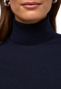 Navy-rippierter Rollkragenoberteil mit einer eng anliegenden Silhouette, das eine glatte Textur und ein hohes Kragen-Design aufweist. Sichtbare silberne Ohringe.