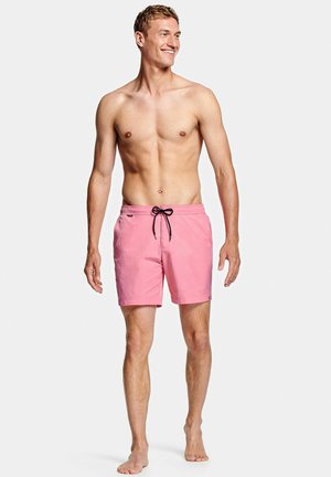 Glimlachende man zonder shirt, gekleed in roze zwemshorts met zwart trekkoord, staat blootsvoets tegen een effen lichte achtergrond.