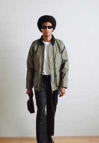 Chaqueta acolchada verde con cuello negro, camiseta blanca, pantalones negros y gafas de sol. Sosteniendo una bolsa negra, el fondo es liso y claro.